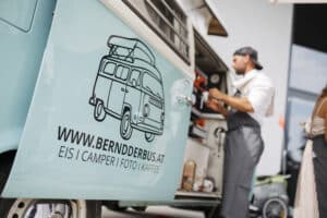 Ein Mann in einer Schürze steht neben einem hellblauen Vintage-Wohnmobil, das mit einem Logo eines Wohnmobils und einer Website-URL geschmückt ist. Der Van ist perfekt für jede Hochzeit und bietet Eiscreme, Wohnmobilservice, Fotografie und Kaffee, um Ihren besonderen Tag unvergesslich zu machen.