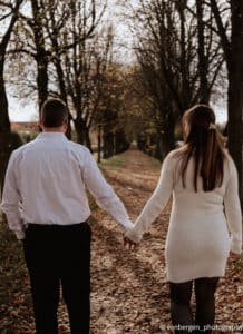 Ein Paar, das seine Hochzeit plant, spaziert Hand in Hand einen laubbedeckten Weg entlang, der von hohen, kahlen Bäumen gesäumt ist. Der Mann trägt ein weißes Hemd und eine schwarze Hose, während die Frau ein cremefarbenes Pulloverkleid mit schwarzen Strumpfhosen trägt. Die Szene ist heiter und herbstlich.