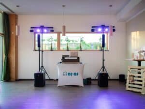 Ein DJ-Setup in einem Raum mit einem Tisch, der mit einem weißen Tuch mit der Aufschrift „DJ GS“ bedeckt ist, sorgt für die perfekte Stimmung bei Ihrer Hochzeit. Der Raum ist mit Lautsprechern, einem Laptop und Beleuchtungsgeräten ausgestattet, elegant mit Holzakzenten dekoriert und große Fenster lassen natürliches Licht herein – ideal für jede Hochzeitsplanung.