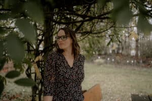Eine Person mit Brille und geblümtem Kleid steht inmitten üppigen Grüns und verkörpert die Gelassenheit einer Hochzeit im Freien. Im Hintergrund sind verschwommene Bäume und ein Rasen zu sehen, die die perfekte Szene für eine Hochzeit inmitten der Schönheit der Natur bilden.