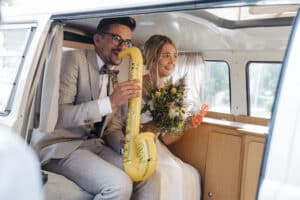Braut und Bräutigam sitzen in einem Oldtimer-Van. Der Bräutigam, in grauem Anzug und Brille, hält verspielt ein gelbes aufblasbares Saxophon. Die Braut, in Weiß gekleidet, lächelt und hält Wildblumen in den Händen. Beide sind glücklich und entspannt an diesem wunderschönen Hochzeitstag.