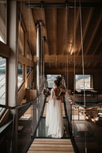 Eine Braut in einem weißen Kleid steht auf einem modernen, schwebenden Steg in einem rustikalen Gebäude mit großen Fenstern. Sie hält einen Blumenstrauß in der Hand und ist ganz in das Wesen ihrer Hochzeit vertieft, während sie auf die schneebedeckte Berglandschaft blickt, die durch das Glas sichtbar ist.