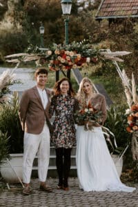 Drei Menschen lächeln unter einem wunderschönen Blumenbogen im Freien und verkörpern die Freude einer Hochzeit. Die Person links trägt eine braune Jacke und weiße Hosen, während die rechte ein weißes Kleid mit einem Blumenstrauß trägt. In der Mitte steht jemand in einem Blumenkleid und vervollständigt diese perfekte Hochzeitsszene.
