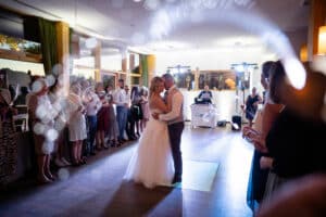 Braut und Bräutigam tanzen ihren ersten Tanz in einem warm beleuchteten Empfangssaal, umgeben von Gästen mit Wunderkerzen in der Hand. Mitten in ihrer Hochzeit umarmt sich das Paar auf der Tanzfläche, während im Hintergrund ein DJ auflegt. Romantisches Ambiente mit festlicher Beleuchtung.