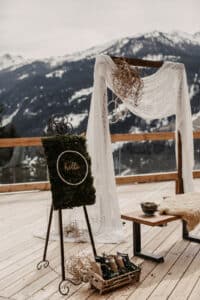 Eine rustikale Hochzeitsdekoration auf einer Holzterrasse mit Blick auf schneebedeckte Berge. Ein Holzbogen ist mit Spitzenstoff behangen, perfekt für alle, die gerade mit der Hochzeitsplanung beschäftigt sind. In der Nähe steht ein mit Moos bedecktes dekoratives Brett mit einem „Hallo“-Schild, während ein Korb mit kleinen Gegenständen auf dem Boden steht.