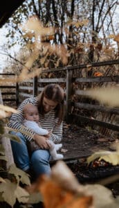 Eine Frau in gestreiftem Hemd sitzt draußen auf einer Holzbank und wiegt ein Baby. Umrahmt von Herbstblättern strahlt die Szene einen warmen, rustikalen Charme aus, der an eine unvergessliche Hochzeit erinnert. Sie lächelt das Baby an, das in die Kamera blickt und sich vielleicht zukünftige Hochzeitstage vorstellt.