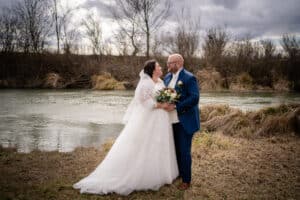 Braut und Bräutigam stehen an einem ruhigen Flussufer, umgeben von kahlen Bäumen unter einem bewölkten Himmel. Die Braut in ihrem weißen Kleid und Schleier mit einem Blumenstrauß in der Hand verkörpert Eleganz. Als Teil ihrer wohlüberlegten Hochzeitsplanung trägt der Bräutigam einen blauen Anzug. Sie blicken einander in die Augen und lächeln herzlich.