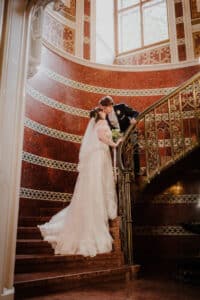 Ein Paar in Hochzeitskleidung steht auf einer verzierten Wendeltreppe und verkörpert einen perfekten Hochzeitsmoment. Die Braut im weißen Kleid hält einen Blumenstrauß, während sich der Bräutigam im schwarzen Anzug zu ihr beugt. Die Treppe verfügt über aufwendige Geländer und reich verzierte Wände, die die Essenz der Hochzeitseleganz einfangen.