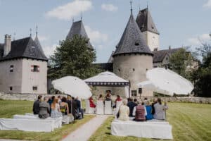 Die Leute sitzen auf Bänken unter Sonnenschirmen und nehmen an einer Hochzeitszeremonie im Freien in einem historischen Schloss mit hohen Türmen und einem Innenhof teil. Der Himmel ist klar mit ein paar Wolken und bietet die perfekte Kulisse für eine Hochzeit in solch einer romantischen Umgebung.