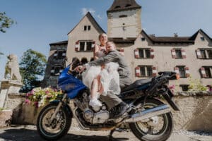 Braut und Bräutigam sitzen mitten in ihrer Hochzeitsplanung auf einem blauen Motorrad vor einem historischen Schloss mit einem hohen Turm. Die Braut trägt ein weißes Kleid und hält ihren Blumenstrauß, während der Bräutigam einen grauen Anzug trägt. Blumen und eine Steinstatue betonen die Szene unter einem klaren blauen Himmel.