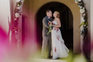 Ein Paar in Hochzeitskleidung umarmt sich unter einem mit weißen und rosa Blumen geschmückten Torbogen und fängt die Essenz einer verträumten Hochzeit ein. Die Braut hält einen Blumenstrauß und trägt ein weißes Kleid mit roten Blumendetails, während der Bräutigam einen grauen Anzug trägt. Der unscharfe Vordergrund trägt zu dieser romantischen Hochzeitsszene bei.