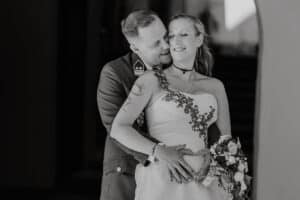 Ein Paar umarmt sich an seinem Hochzeitstag. Der Bräutigam in Militäruniform hält die Braut, die ein weißes Kleid mit Blumendetails trägt und einen Blumenstrauß hält. Der Bräutigam formt mit seinen Händen ein Herz über dem Bauch der Braut. Diese Hochzeitsszene ist fröhlich und intim.