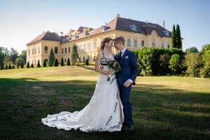 Braut und Bräutigam stehen gemeinsam vor einem großen, eleganten Gebäude, möglicherweise einem Herrenhaus oder Schloss. Die Braut trägt ein weißes Kleid mit schwarzer Stickerei und hält einen Blumenstrauß, während der Bräutigam einen blauen Anzug trägt. Sie feiern ihre Hochzeit in einem sonnenbeschienenen Garten.