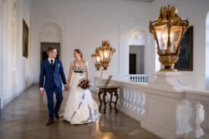 Ein Paar in Abendgarderobe geht Hand in Hand durch einen eleganten Flur mit verzierten Lampen und weißen Wänden und verkörpert den Geist ihrer Hochzeit. Die Frau trägt ein weißes Kleid mit schwarzen Akzenten und hält einen Blumenstrauß, während der Mann einen blauen Anzug trägt. Sie lächeln sich an, verloren in dem Moment.