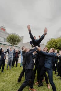 Eine Gruppe von Menschen in Anzügen hebt freudig eine Person auf einer Rasenfläche in die Luft und feiert eine Hochzeit. Im Hintergrund sind ein Gebäude und einige Bäume zu sehen, darüber ein bewölkter Himmel. Die Stimmung ist festlich und erfüllt von Hochzeitsfreude.