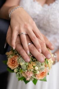 Nahaufnahme der Hände eines Paares mit Eheringen über einem Strauß pfirsichfarbener und weißer Blumen, die die Essenz ihrer Hochzeit einfangen. Die Braut trägt ein Spitzenkleid und ein silbernes Armband, perfekte Details in ihrer Traumhochzeitsplanung.