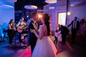 Braut und Bräutigam tanzen bei ihrem Hochzeitsempfang gemeinsam, in sanftes Licht getaucht, während im Hintergrund eine Live-Band spielt. Gäste, darunter ein Mann, der einem Kind hilft, schwelgen in der festlichen Atmosphäre und fangen die Magie dieser wunderschön geplanten Feier ein.