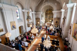 Luftaufnahme des Kircheninneren während einer Hochzeitszeremonie. Die Menschen sitzen in Holzbänken, einige stehen. Der Altar, perfekt für die Hochzeitsplanung, ist mit religiösen Gemälden geschmückt und Kronleuchter hängen von der Decke. Die Architektur zeichnet sich durch hohe Bögen und kunstvolle Verzierungen aus.