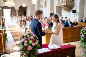 Inmitten des bezaubernden Sonnenlichts, das durch die Buntglasfenster fällt, tauschen Braut und Bräutigam in einer wunderschön geschmückten Kirche die Ringe aus. Die sorgfältige Hochzeitsplanung ist in jedem Detail erkennbar. Die Gäste sitzen in Holzbänken und erleben diesen herzlichen Moment der Hochzeit umgeben von eleganten Blumen.