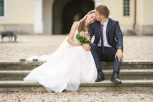 Ein frisch vermähltes Paar sitzt draußen auf Steinstufen, während der Bräutigam den Kopf der Braut küsst. In diesem intimen Moment ihrer Hochzeit hält sie einen Strauß rosa und grüner Blumen in der Hand und legt ihren Kopf auf seine Schulter. Der Bräutigam trägt einen marineblauen Anzug, während die Braut ein fließendes weißes Kleid trägt.