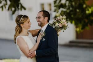 Braut und Bräutigam stehen draußen und lächeln sich an ihrem Hochzeitstag an. Die Braut hält einen Strauß rosafarbener und weißer Blumen in der Hand und trägt ein weißes Kleid mit einem schlichten Haarband. Der Bräutigam trägt einen dunklen Anzug mit einer zum Strauß passenden Ansteckblume. Im Hintergrund sind Bäume zu sehen.