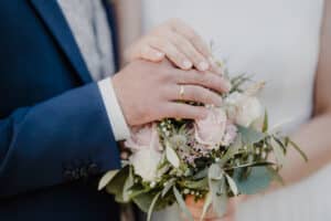 Braut und Bräutigam halten sich an den Händen, ihre Eheringe glänzen über einem Strauß rosa und weißer Blumen mit üppigem Grün. Der dunkelblaue Anzug des Bräutigams ergänzt die Kulisse perfekt, während die weiß lackierten Nägel der Braut ihrem Hochzeitsmoment einen Hauch von Eleganz verleihen.
