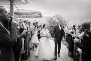Ein Schwarzweißfoto fängt eine Hochzeitsszene ein, in der Braut und Bräutigam lächeln, während sie durch eine Menge Gäste gehen und Seifenblasen pusten. Die Braut in einem trägerlosen Kleid mit einem Blumenstrauß in der Hand und der Bräutigam im Anzug sorgen auf dem überdachten Außenweg für eine festliche Atmosphäre.