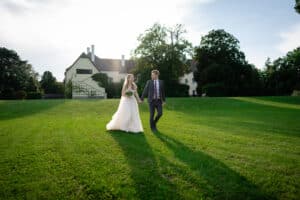 Braut und Bräutigam gehen an ihrem Hochzeitstag Hand in Hand über einen weitläufigen, üppig grünen Rasen. Die Braut trägt ein weißes Kleid und der Bräutigam einen Anzug. Hinter ihnen steht ein großes Haus, umgeben von Bäumen unter einem klaren, sonnigen Himmel.
