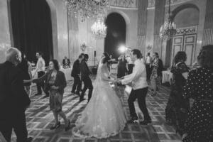 Ein Schwarzweißfoto fängt eine Hochzeitsszene in einem eleganten Ballsaal ein. Ein Paar in Hochzeitskleidung tanzt anmutig in der Mitte, umgeben von anderen Gästen. Der Raum, perfekt zum Heiraten, verfügt über Kronleuchter, hohe Vorhänge und kunstvolle Details, während formell gekleidete Gäste zuschauen.