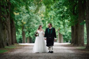 Eine Braut im weißen Kleid und ein Bräutigam im schwarzen Anzug gehen Hand in Hand einen von Bäumen gesäumten Weg entlang und verkörpern damit die Essenz einer Hochzeit. Umgeben von üppigen grünen Bäumen feiern sie ihre Liebe in einer ruhigen und romantischen Atmosphäre.