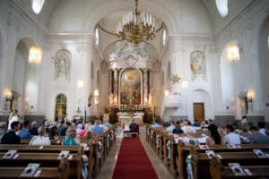 Der Innenraum der Kirche ist für eine Hochzeit hergerichtet. Die Gäste sitzen in den Bänken vor dem Altar, der für diese wunderschöne Hochzeitszeremonie geschmückt ist. Ein roter Teppich ziert den Mittelgang, der zu kunstvoller Architektur, Kronleuchtern und hohen Decken führt – eine perfekte Szene für Hochzeiten.