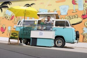 Ein Mann serviert Eiscreme aus einem türkis-weißen Oldtimer-Van, der vor einem lebendigen Wandgemälde geparkt ist, das sich perfekt als skurriler Hochzeitshintergrund eignet. Das Set-up umfasst eine Glasvitrine, ein Schild und einen gelben Regenschirm. Das Wandgemälde mit cartoonartigen Figuren und leuchtenden Farben verleiht der Hochzeitsplanungsszene Charme.
