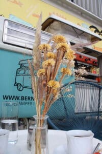 Ein Strauß getrockneter Blumen in einer Vase ziert den Tisch im Freien und deutet auf eine durchdachte Hochzeitsplanung hin. Im Hintergrund zeigt ein hellblauer Campervan einen Vintage-Touch, während zwei Tassen Kaffee und Gläser Wasser eine ruhige Flucht vor den Hochzeitsfeierlichkeiten vorschlagen.
