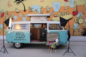 Ein hellblauer Oldtimer-Van mit offener Tür ist mit Stühlen und Requisiten wunderschön für ein Hochzeitsplanungs-Fotoshooting hergerichtet. Er steht vor einem farbenfrohen Wandgemälde im Cartoon-Stil. Auf beiden Seiten des Vans sind zwei Fotolichter angebracht.