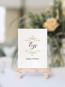 Eine Hochzeitseinladungskarte mit eleganter Kalligraphie in Gold und Schwarz, mit den Initialen „E & P“ über den Namen „Esther & Peter“, perfekt für Ihre Hochzeit. Die Karte wird auf einer kleinen Holzstaffelei mit verschwommenem Blumenhintergrund präsentiert, ideal für diejenigen, die schon weit in ihrer Hochzeitsplanung stecken.