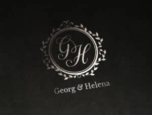 Ein silbernes geprägtes Logo auf dunklem Hintergrund zeigt die Initialen „G“ und „H“ in einem kunstvollen Kreisdesign, das mit dekorativen Schnörkeln geschmückt ist. Unter dem Kreis ist der Text „Georg & Helena Hochzeitsplanung“ in einer klassischen Serifenschrift gedruckt.
