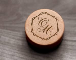 Ein runder Deckel einer Holzbox mit den Initialen „GM“ in eleganter Schrift, umgeben von einem geometrischen Rand, ist perfekt für die Hochzeitsplanung. Er ruht wunderschön auf einer grauen strukturierten Oberfläche und verleiht Ihrem besonderen Tag Charme.