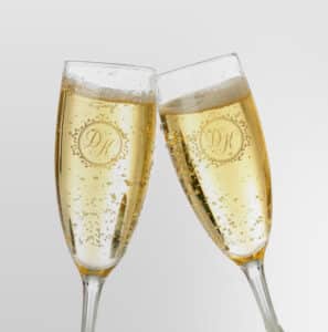 Zwei Champagnergläser berühren sich leicht, gefüllt mit prickelndem Champagner – ein Bild des Eheglücks. Die Gläser sind mit einem dekorativen Monogramm „PK“ in einem kreisförmigen Design eingraviert, das den Geist der Hochzeit perfekt einfängt. Der schlichte Hintergrund unterstreicht die feierliche Stimmung.