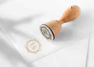 Ein Holzstempel mit dem Monogramm „WM“ liegt auf einem weißen Umschlag und hinterlässt seinen Abdruck auf dem Papier. Perfekt für die Hochzeitsvorbereitungen: Das Monogramm ist von einem dekorativen runden Rand umgeben, der jeder Hochzeitseinladung eine elegante Note verleiht.
