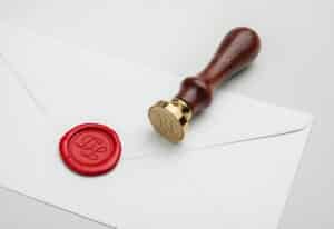 Ein weißer Umschlag mit einem roten Wachssiegel ist sichtbar, perfekt für Ihre Hochzeitsplanung. Daneben liegt ein Siegelstempel mit einem Holzgriff und einer goldenen gravierten Platte. Das gravierte Design auf dem Stempel entspricht dem Design auf dem Wachssiegel und verleiht jeder Hochzeitseinladung eine elegante Note.