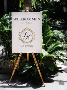 Auf einem Hochzeits-Willkommensschild auf einer Staffelei, umgeben von üppigem Grün, steht „WILLKOMMEN zur Hochzeit“ mit einem dekorativen Monogramm „LK“ und den Namen „Lisa & Konrad“ und gibt damit den Ton für ihre sorgfältig ausgearbeitete Hochzeitsplanung an.