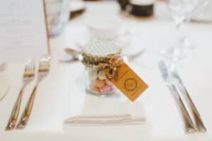 Ein kleines Glasgefäß mit gepunktetem Stoffdeckel enthält verschiedene Süßigkeiten, perfekt für die Feier einer Hochzeit. Mit einer Schleife zusammengebunden und mit einem Dankeschön-Etikett versehen, steht das Glas elegant auf einem hübsch gedeckten Esstisch neben Besteck und Gläsern und ist bereit, Hochzeitsgäste zu erfreuen.