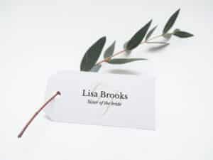 Ein weißes Etikett mit dem Text „Lisa Brooks, Schwester der Braut“ in eleganter Goldschrift ist an einem schlanken Eukalyptuszweig mit grünen Blättern auf weißem Hintergrund befestigt, perfekt für Ihre Hochzeitsdekoration.