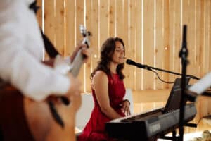 In einem warm beleuchteten, rustikalen Veranstaltungsort mit Holztäfelung singt und spielt eine Frau in einem roten Kleid Keyboard und schafft so eine bezaubernde Atmosphäre, die perfekt für Ihre Hochzeit ist. Ihre gefühlvolle Melodie wird wunderbar von einem Gitarristen ergänzt und macht es zu einem idealen Ort, um nach einem Tag voller Hochzeitsplanung zu entspannen.