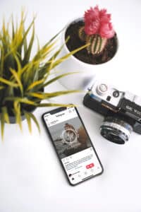 Ein Smartphone, auf dem ein Instagram-Beitrag angezeigt wird, liegt auf einer weißen Oberfläche neben einer Vintage-Kamera, einer stacheligen Zimmerpflanze im Topf und einem kleinen Kaktus in einem weißen Topf – so fängt jedes Detail Inspiration für die Hochzeitsplanung ein.