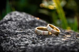 Zwei goldene Eheringe mit eingravierten Buchstaben liegen auf einem strukturierten grauen Stein und symbolisieren die Reise der Heiraten. Der Hintergrund ist sanft verschwommen und deutet auf grünes Laub hin, wodurch eine ruhige Außenkulisse entsteht, die sich perfekt für die Hochzeitsplanung eignet.