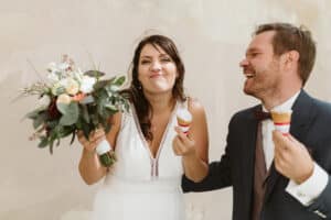 Braut und Bräutigam halten fröhlich Eistüten in den Händen und verkörpern damit die lustige Seite der Hochzeitsplanung. Die Braut, in einem weißen Kleid, hält ebenfalls einen Blumenstrauß. Beide lächeln und lachen in einem unbeschwerten Moment vor dem neutralen Wandhintergrund.