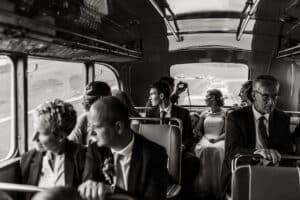 Ein Schwarzweißfoto fängt einen heiteren Moment während einer Hochzeit ein: Braut und Bräutigam inmitten von Passagieren in Abendgarderobe in einem Bus, die aus dem Fenster oder einander ansehen.