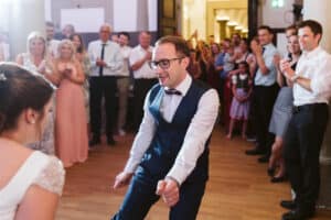 Ein Mann in Weste und Fliege tanzt begeistert auf einem Hochzeitsempfang, umgeben von applaudierenden Gästen. Ihm gegenüber steht eine Frau im Brautkleid, die ebenfalls tanzt. Die Atmosphäre ist fröhlich und feierlich.