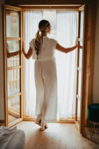 Eine Frau in weißem Outfit steht vor offenen Holztüren und blickt auf eine helle Szene, die an eine Hochzeit erinnert. Sie hält den Türrahmen mit den Händen fest, ihr langer Zopf fällt anmutig herab. Der Raum verfügt über Holzboden und einen grünen Hocker an der Seite, perfekt für die Hochzeitsplanung.